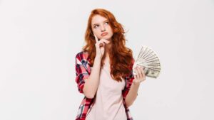 Mulher ruiva segurando cédulas de dinheiro, aparentando estar indecisa sobre o que fazer. A imagem faz alusão ao texto ensinando a como lidar com dinheiro.